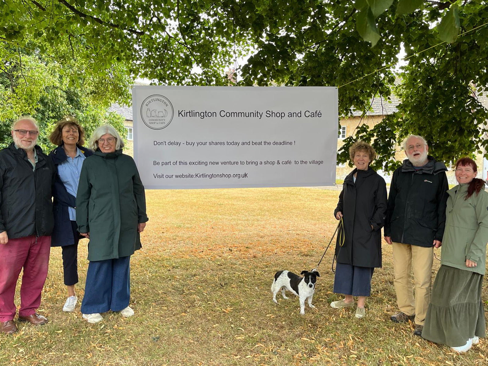 Group of people with a dog standing in front of a sign for Kirlington Community Shop and Café.